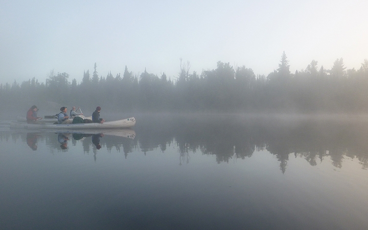 A canoe is paddled by outward bound students on very calm water. The air is foggy, and the water reflects the tree-lined shore. 